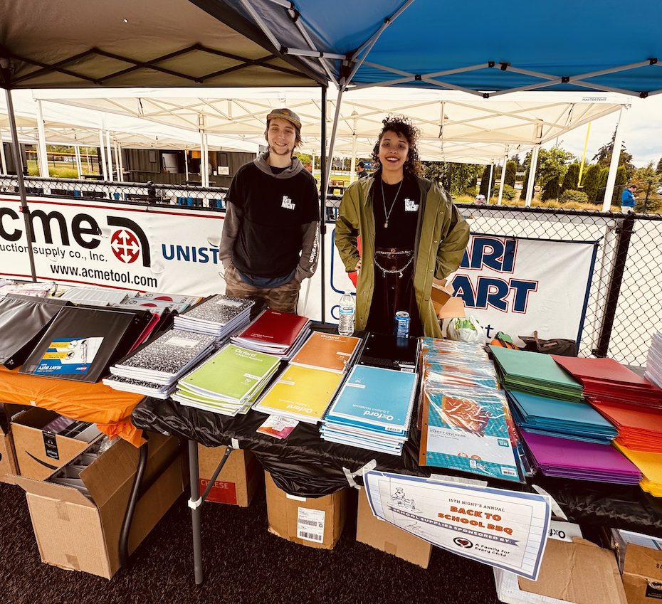 Two members of the youth action council stand side by side behind a table of school supplies. There are folders, binders, spiral notebooks laid across the table.